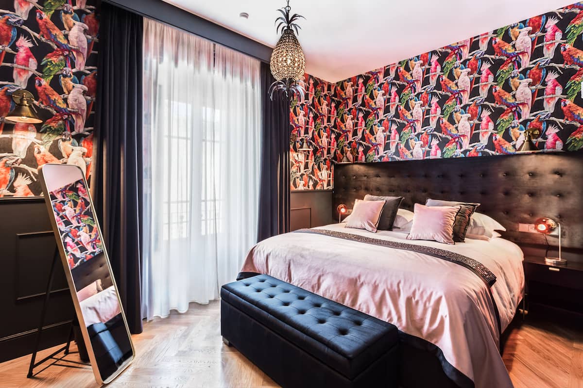 Un dormitorio con una cama grande, un banco tapizado negro, un espejo y una pared con un estampado de pájaros coloridos.