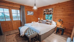 A wooden cabin bedroom with a large bed, a bench, a nightstand, and a view of the outdoors.