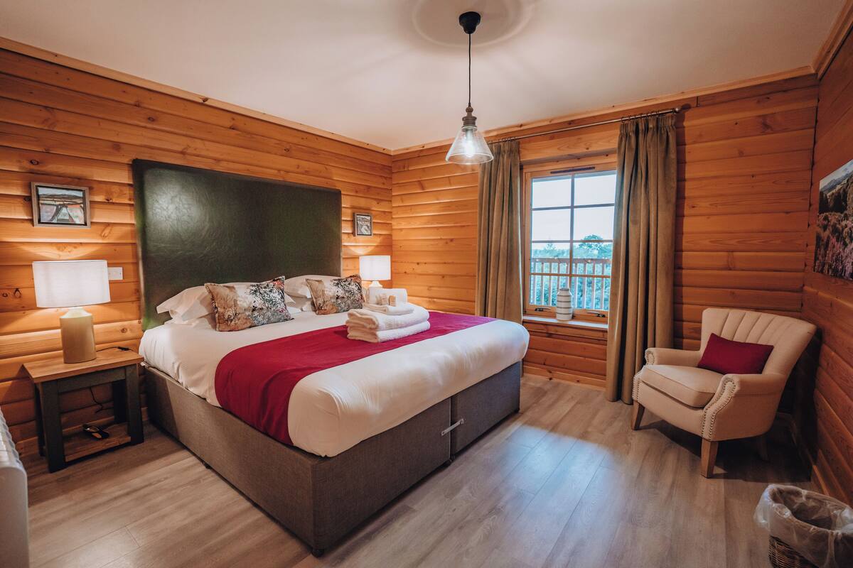 A wooden cabin bedroom with a bed, bedside lamps, a chair, and a window with curtains.