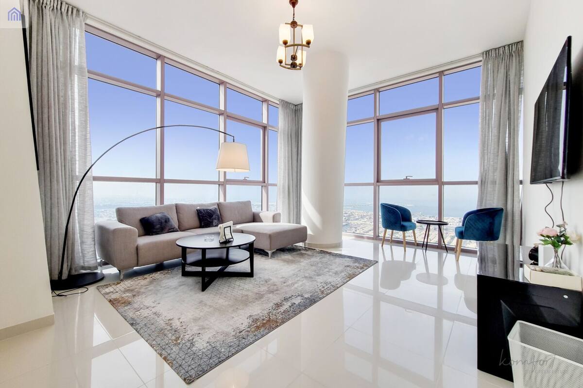 A modern living room with a sofa, coffee table, and chairs, large windows with a city view, and a flat-screen TV.
