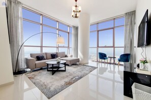 A modern living room with a sofa, coffee table, and chairs, large windows with a city view, and a flat-screen TV. - GreenFuture - A A Tower - Two beedroms (Dubai)