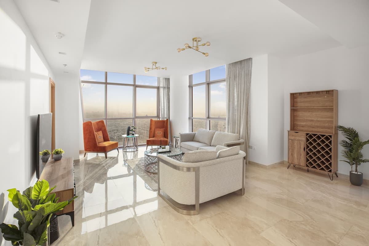 A modern living room with a sofa, armchairs, a coffee table, and a TV. Large windows offer a view of the cityscape.