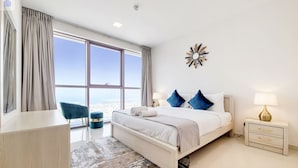 Una habitación de hotel moderna con un ventanal grande, una cama con ropa blanca, un sillón azul, un pequeño reposapiés y una mesita de noche con una lámpara.
