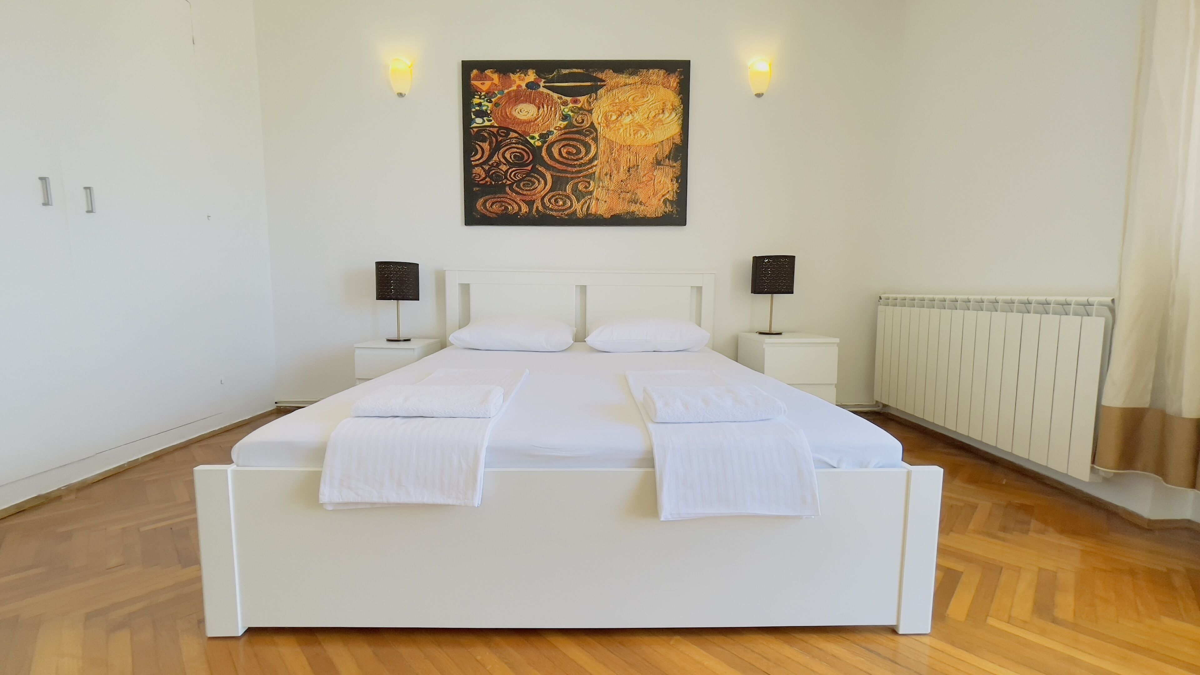 Una cama bien hecha con sábanas y toallas blancas, dos mesitas de noche con lámparas, un radiador y un cuadro abstracto grande en la pared.