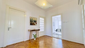 A spacious room with a wooden herringbone floor, white walls, and a ceiling light. There is a door with a handle, a small table with a plant, and a painting on the wall. - Apartment Reful (Kraljevica)
