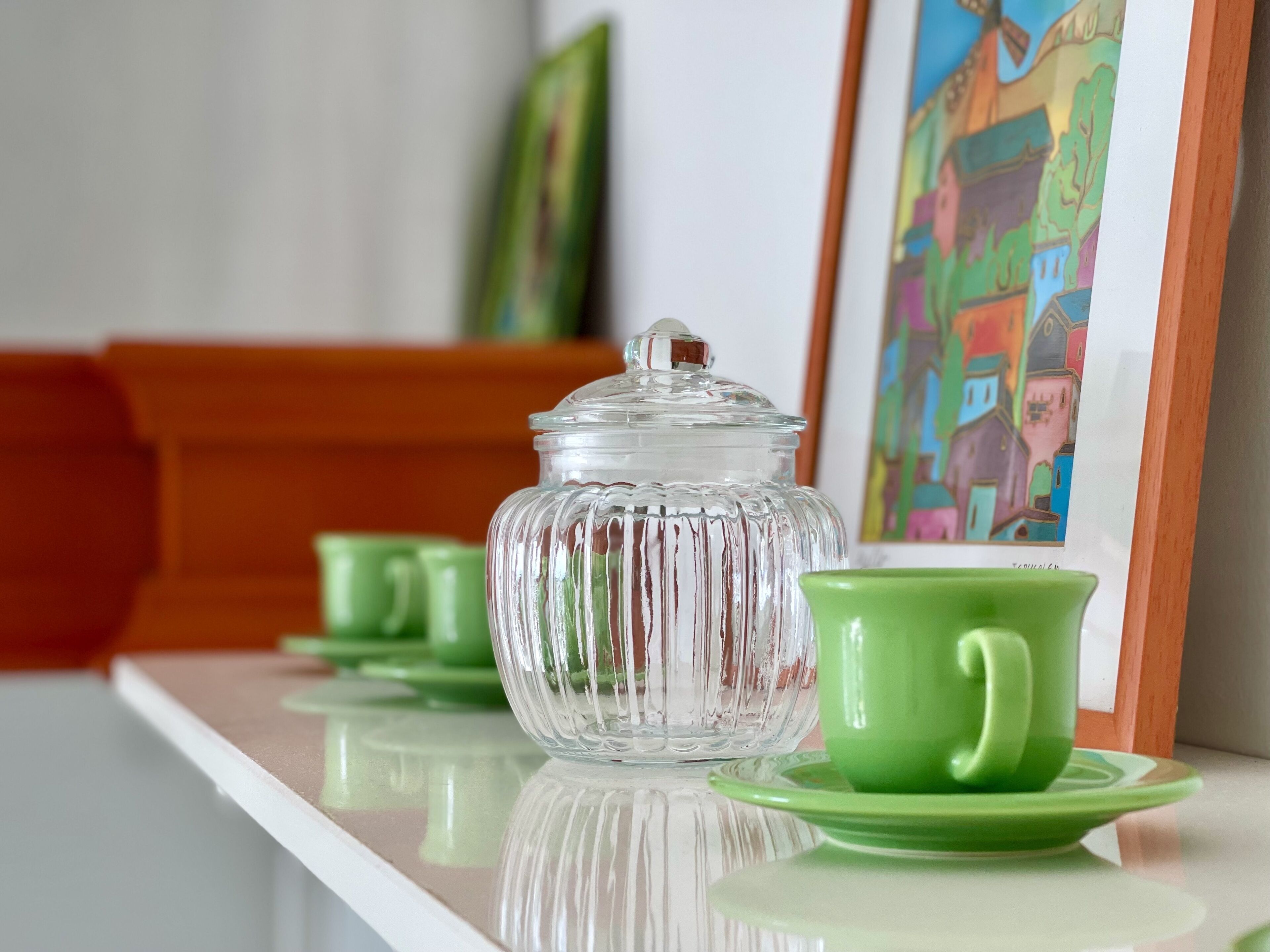 Un frasco de vidrio transparente con tapa sobre una superficie reflectante, junto con una taza y un platillo a juego en color verde.
