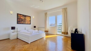 A modern bedroom with a large bed, two bedside tables with lamps, a ceiling light, a large window with curtains, and a view of buildings outside. - Apartment Reful (Kraljevica)