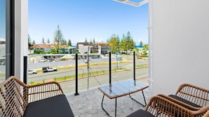Varanda com móveis de vime e uma mesa de vidro, com vista para uma rua com edifícios e árvores.