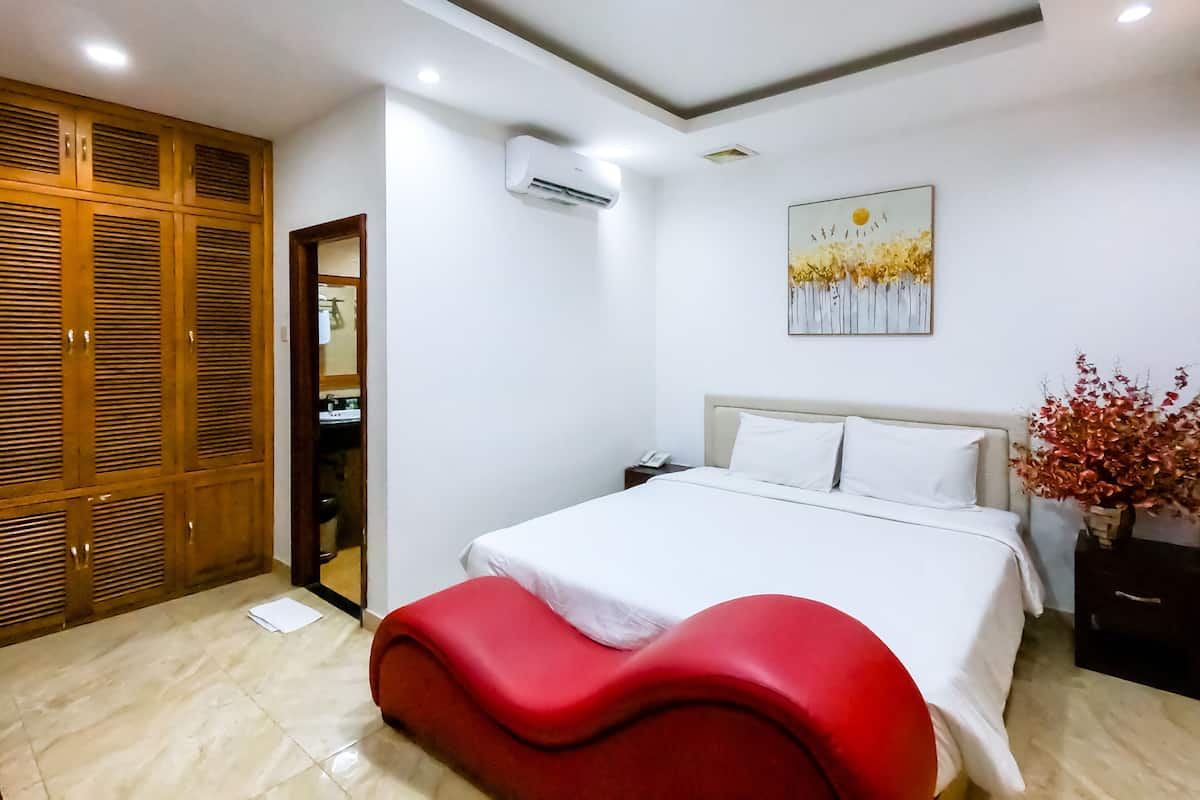 Quarto de hotel com uma cama, um banco vermelho, um guarda-roupa, ar condicionado, um quadro e uma mesinha de cabeceira.