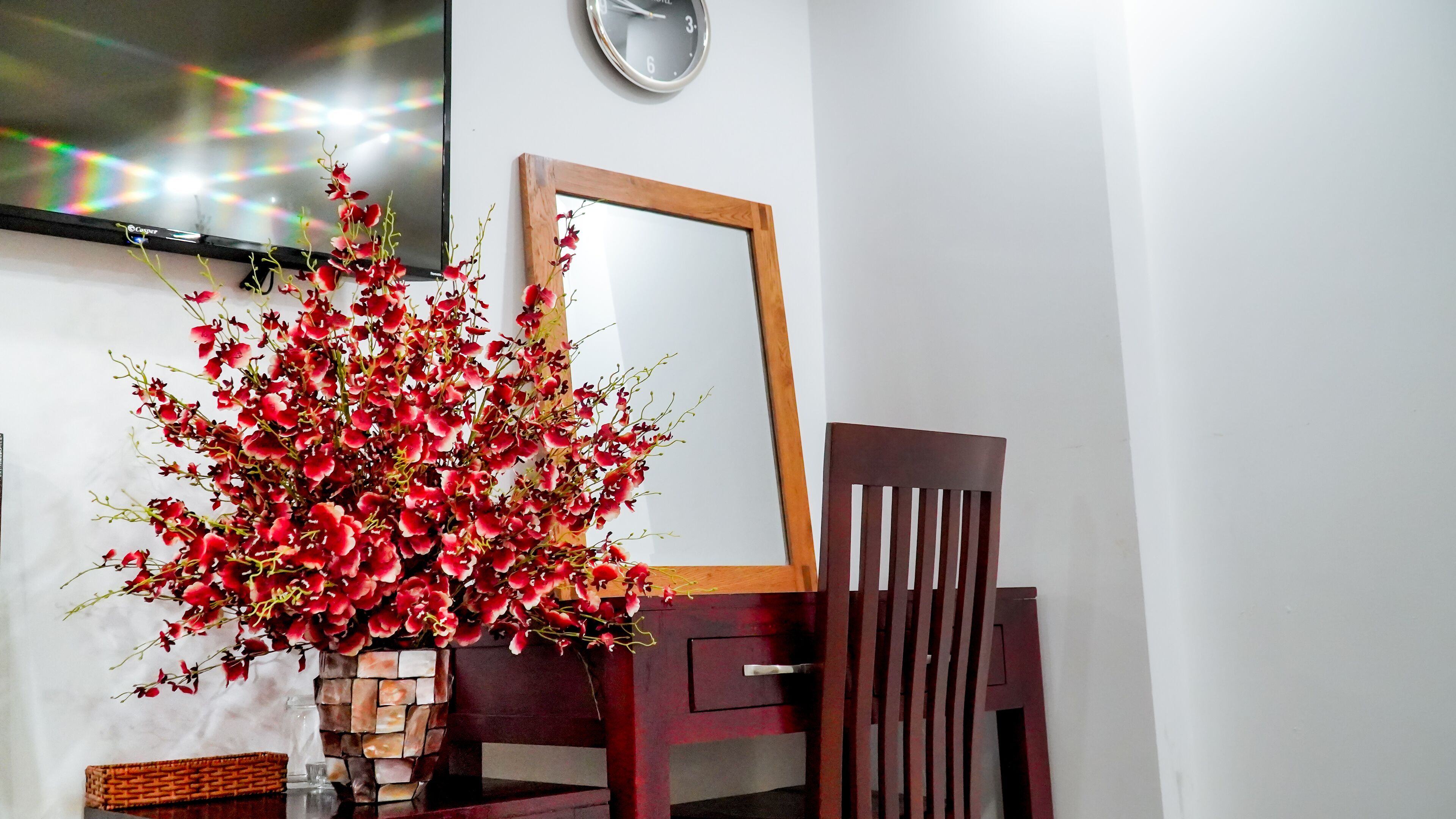 Una habitación con una silla de madera, un espejo y un jarrón con flores rojas sobre una mesa.