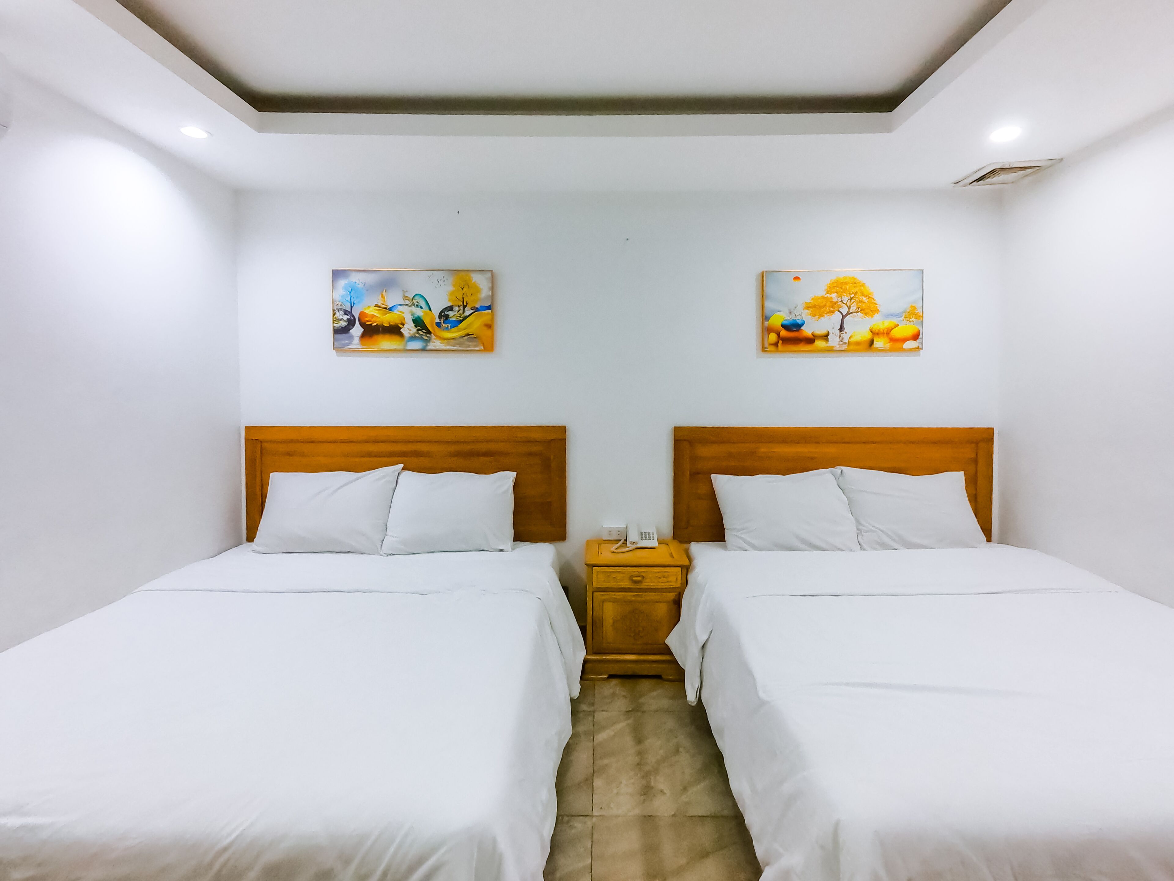 Una camera d'albergo con due letti singoli, poggiatesta in legno, biancheria da letto bianca, un comodino con un telefono e due quadri appesi alle pareti.