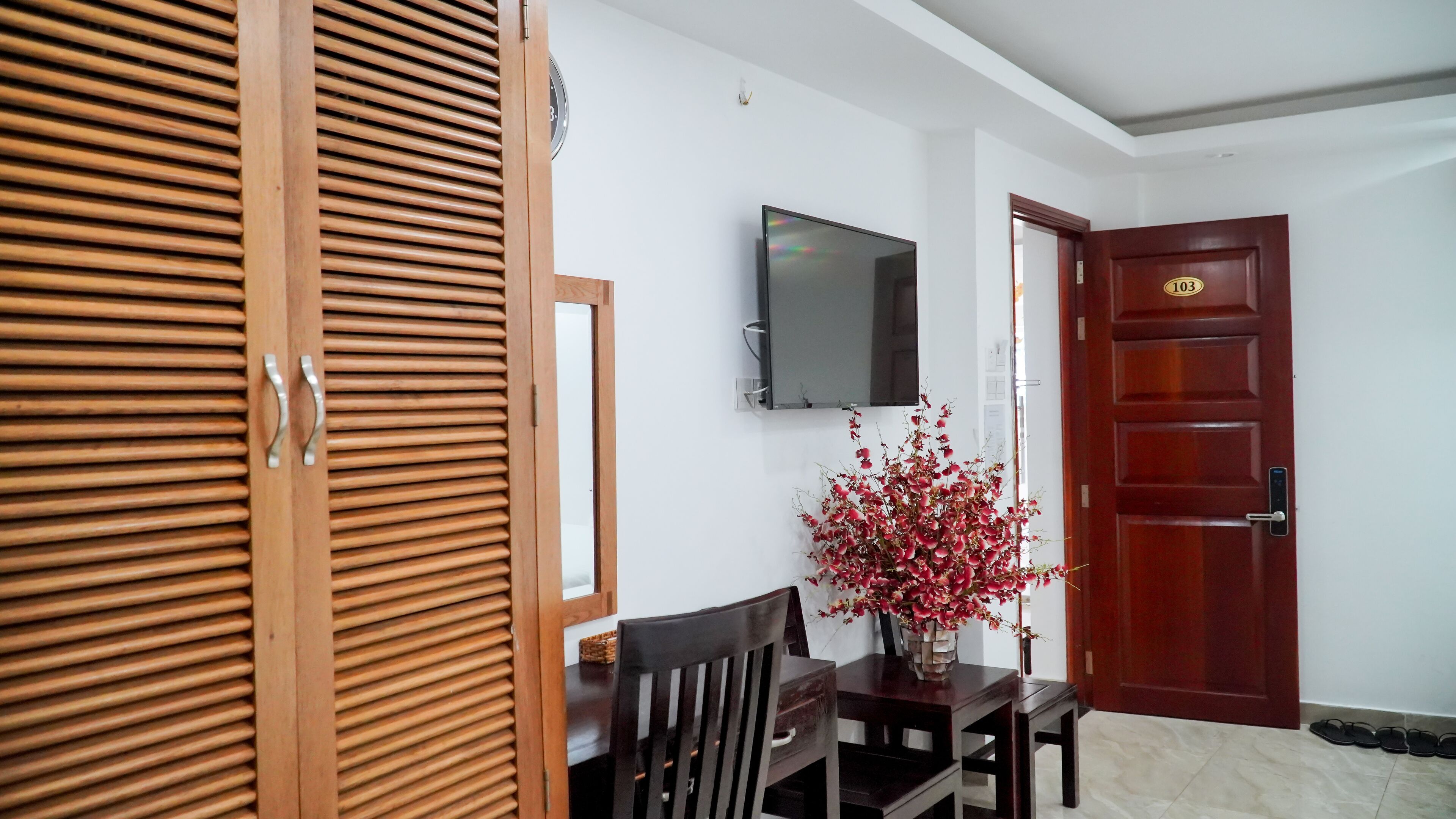 A room with a wooden door, a television mounted on the wall, a dining table with chairs, and a vase with red flowers.