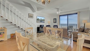 A spacious living area with a glass dining table, a sofa, a TV, and a balcony with a view.