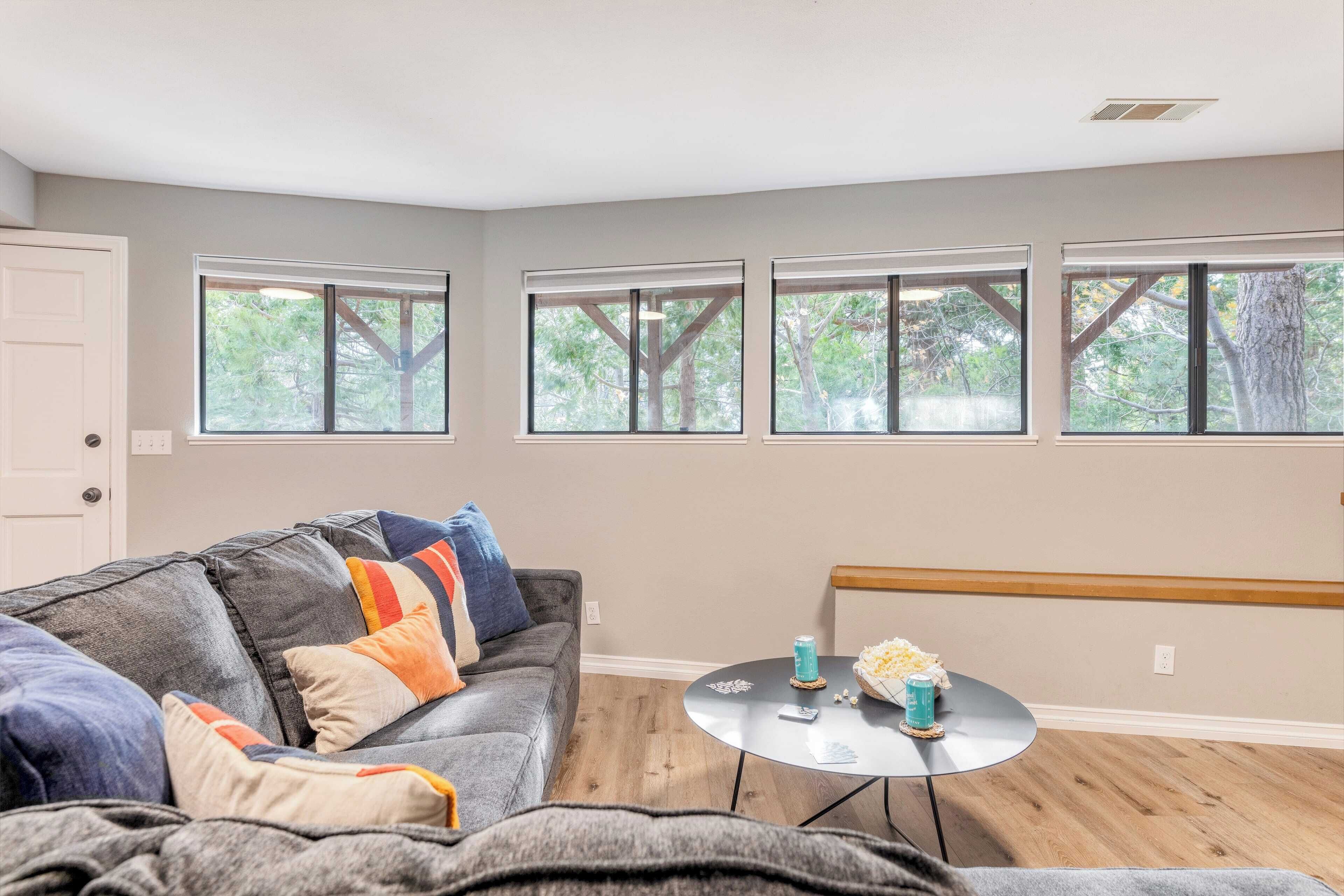 A living room with a gray sectional sofa, a round coffee table, and a view of trees through large windows.