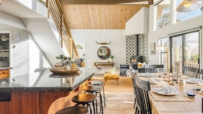 A modern kitchen with a central island, wooden accents, and a dining area with a view. - North Bay by Avantstay Chic Retreat w/ Peloton (Lake Arrowhead)