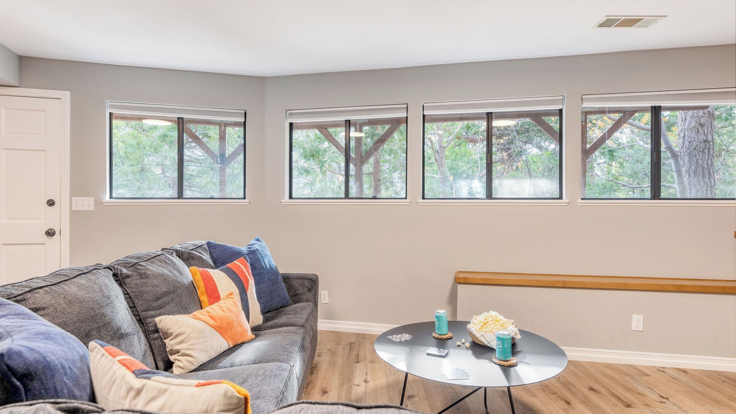 A living room with a grey sofa, three windows with a view of trees, a small round table with drinks, and a wooden bench.