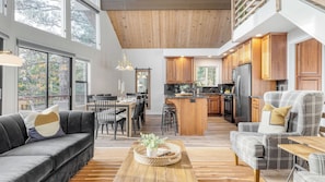 A spacious living area with a sofa, armchair, and coffee table, a kitchen with wooden cabinets and appliances, and a dining area with chairs and a table. - North Bay by Avantstay Chic Retreat w/ Peloton (Lake Arrowhead)