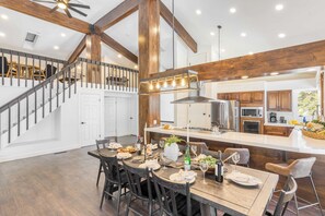 A spacious kitchen with wooden beams, a dining area with a large island, and a staircase leading to an upper level. - Explorer by Avantstay Close to Slopes! Game Room, Guest Suite, Hot Tub (Olympic Valley)