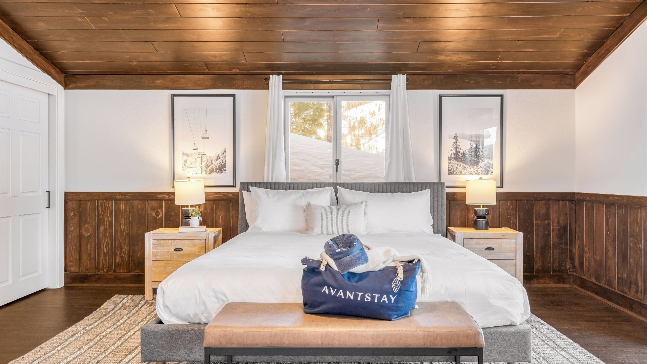Ein Schlafzimmer mit Holzbalkendecke, einem großen Bett mit weißer Bettwäsche, zwei Nachttischen mit Lampen, einem Fenster mit Blick auf Bäume und einer blauen Tasche mit dem Aufdruck 'AVANTSTAY' auf dem Bett.