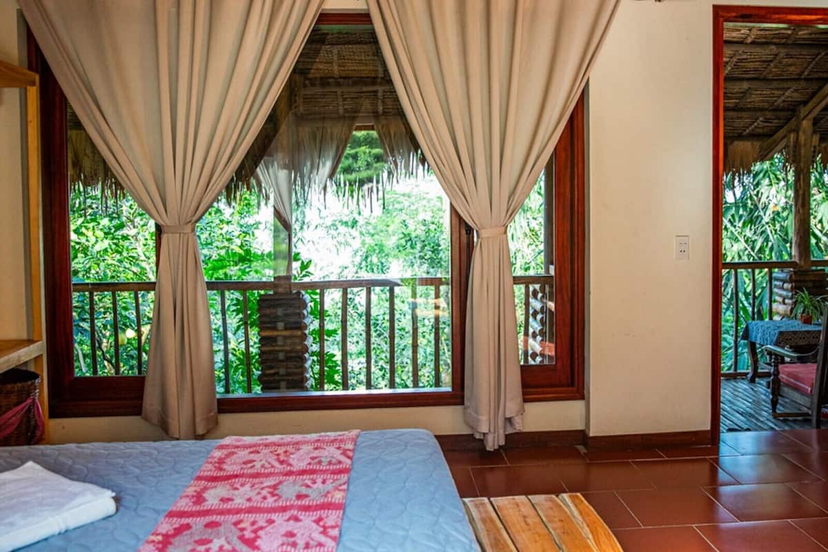 A bedroom with a bed, curtains, a view of greenery, a wooden bench, and a small table with a plant.