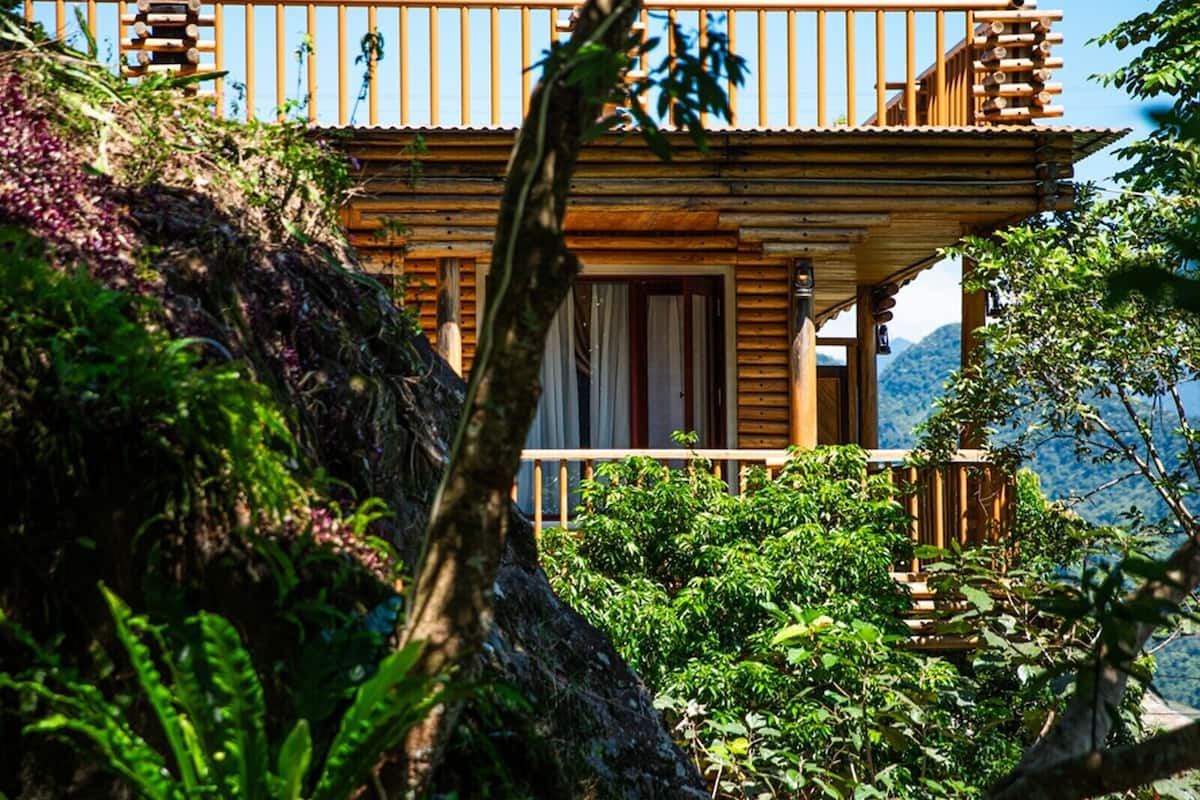 A wooden cabin with a balcony, surrounded by lush greenery and mountains in the background.