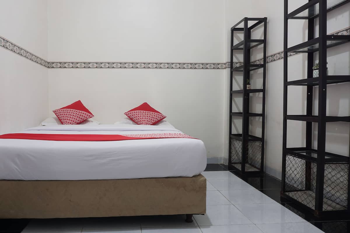 A neatly made bed with white bedding and red pillows, a beige upholstered headboard, and a black metal shelving unit against the wall.