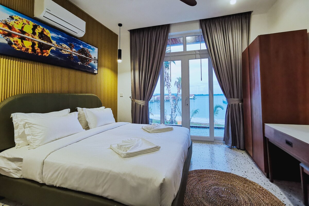 A hotel room with a bed, a desk, a ceiling fan, an air conditioning unit, a painting, and a view of the water outside.