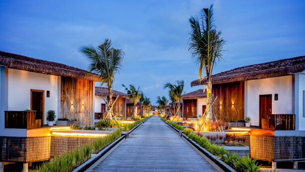 Un resort con un paseo marítimo de madera que conduce a bungalows sobre el agua, cada uno con techo de paja y rodeado de palmeras.