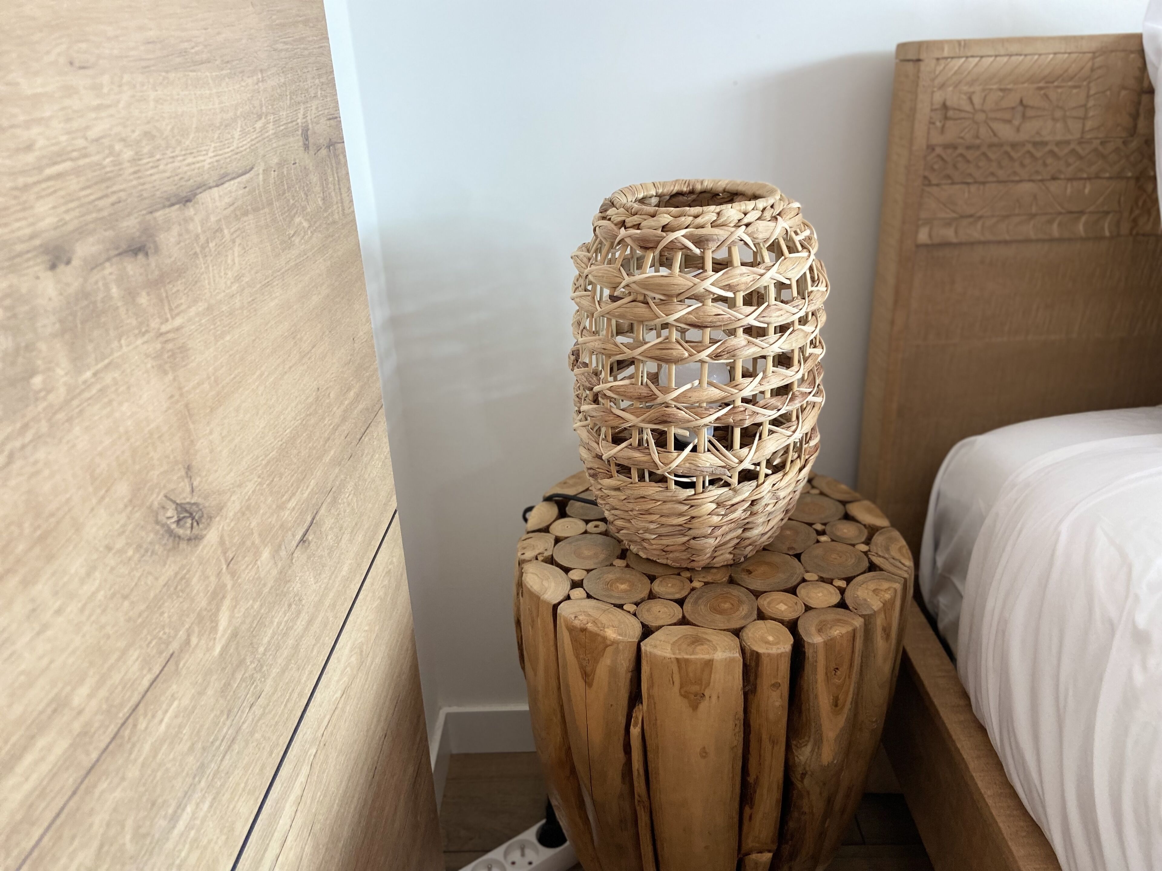A wooden bedside table with a woven basket on top, a wooden headboard, and a white bedspread.