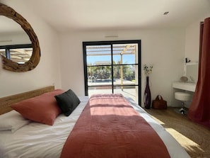 Ein Schlafzimmer mit Bett, großem Fenster, Spiegel, Nachttisch und angrenzendem Badezimmer.