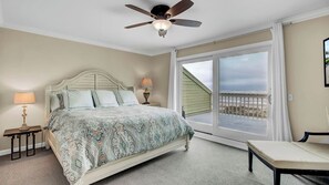 A bedroom with a bed, a ceiling fan, a nightstand with a lamp, a chair, and a view of the ocean through a sliding door.