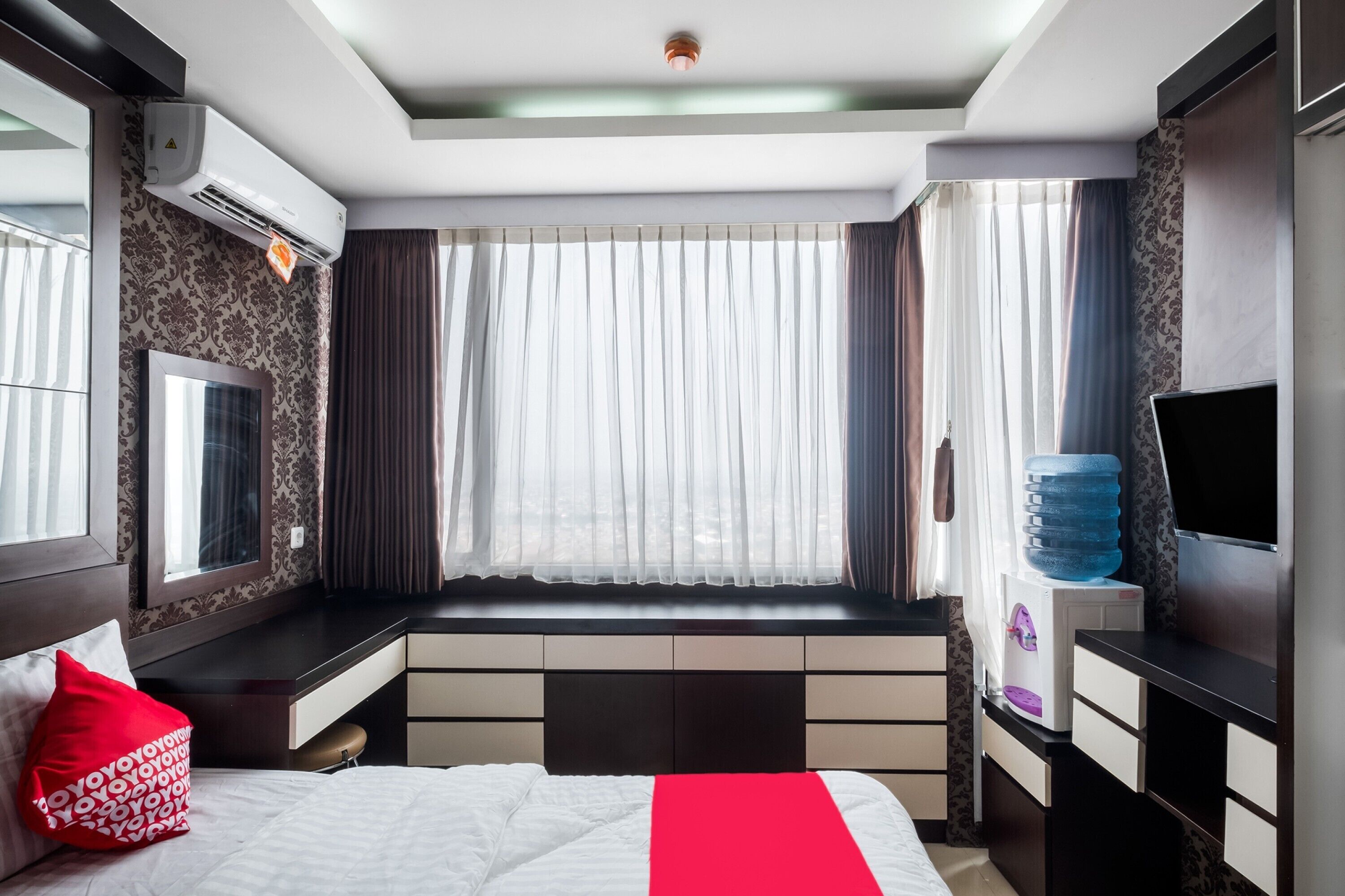 A modern hotel room with a large bed, a built-in desk, a flat-screen TV, a water dispenser, and a window with curtains.