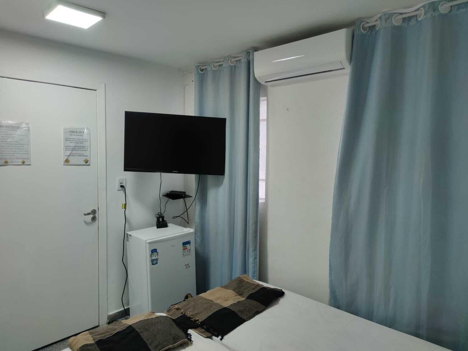 A small, simple room with a bed, a flat-screen TV mounted on the wall, a small white cabinet, and a window with curtains.