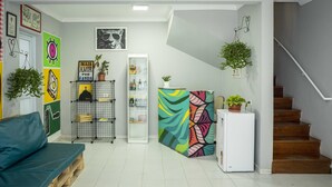 Una sala moderna con un sofá, un refrigerador, una escalera y varias decoraciones en las paredes, incluyendo cuadros enmarcados y un cuadro abstracto colorido.