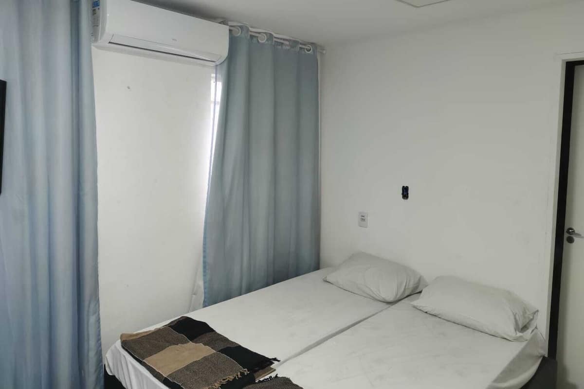 Una piccola camera con un letto singolo, un letto bianco, una finestra con tende azzurre e un condizionatore d'aria bianco a parete.