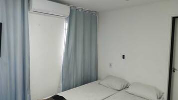 A small, single-bed room with a white bed, a window with light blue curtains, and a white air conditioning unit on the wall.