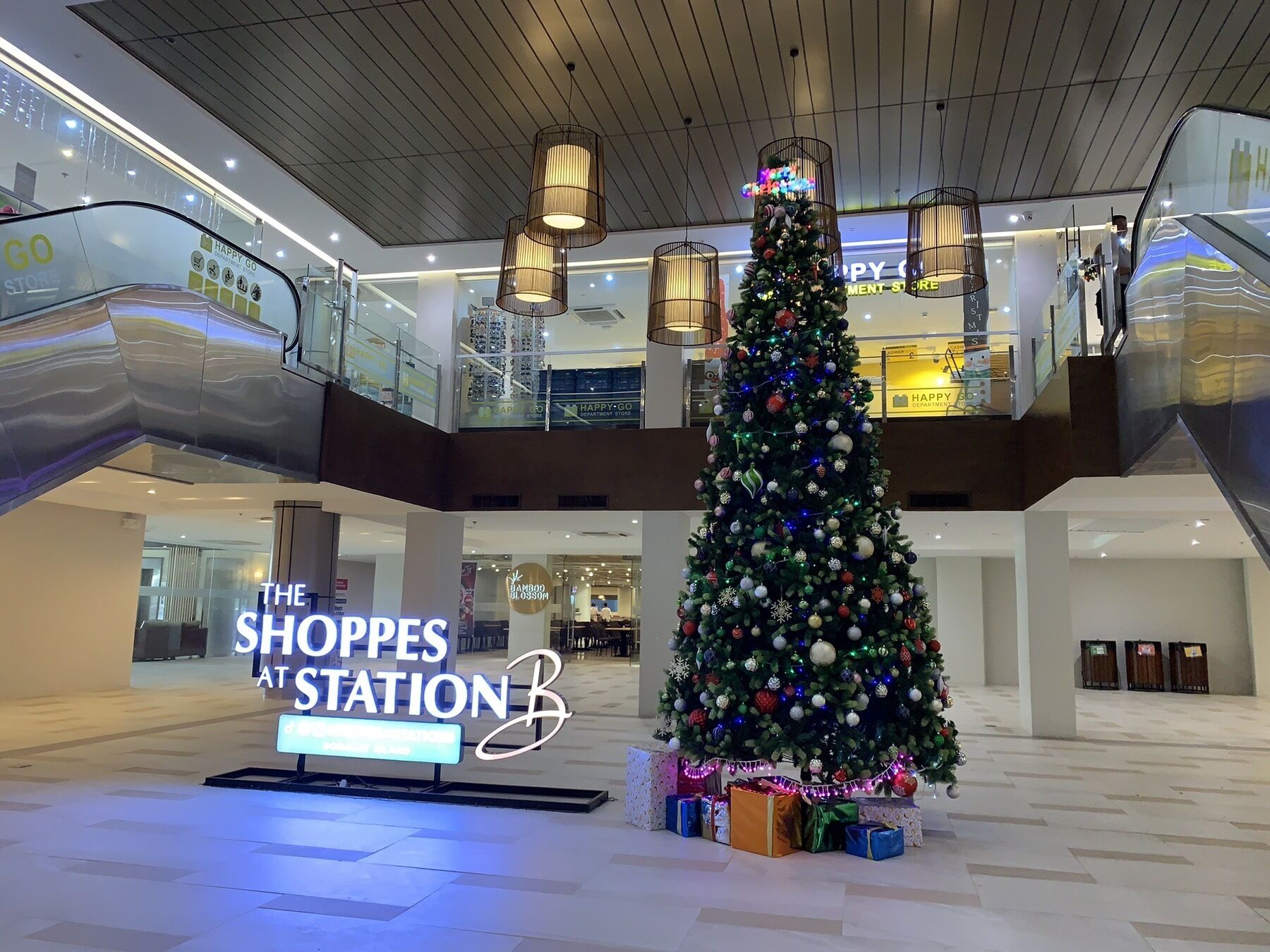 A shopping mall interior with a large Christmas tree, escalators, and a sign for 'The Shops at Station B'.