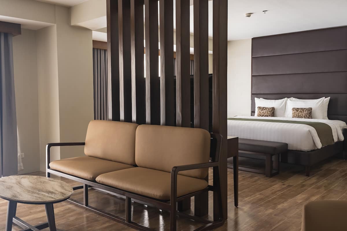 Ein modernes Hotelzimmer mit braunem Ledersofa, einem runden Tisch und einem großen Bett mit dunklem Kopfteil.