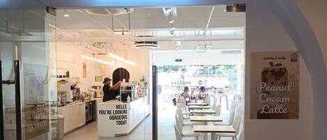 A cafe entrance with a modern design, a digital sign displaying the cafe's name, and a seating area visible inside.