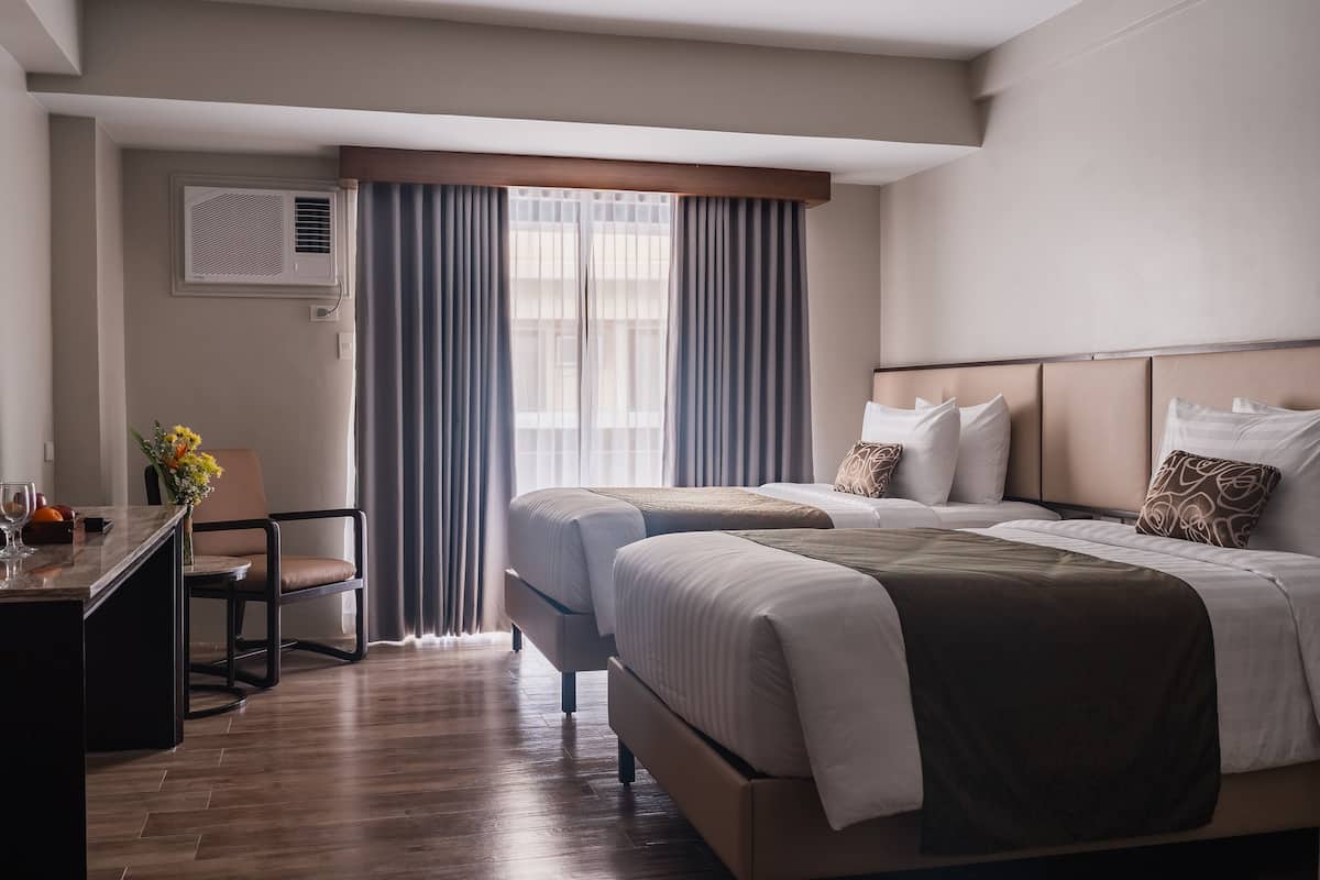 Ein Hotelzimmer mit zwei Betten, einem Schreibtisch und einem Stuhl. Es gibt ein Fenster mit Vorhängen, eine Klimaanlage und einen Blumenstrauß auf dem Schreibtisch.