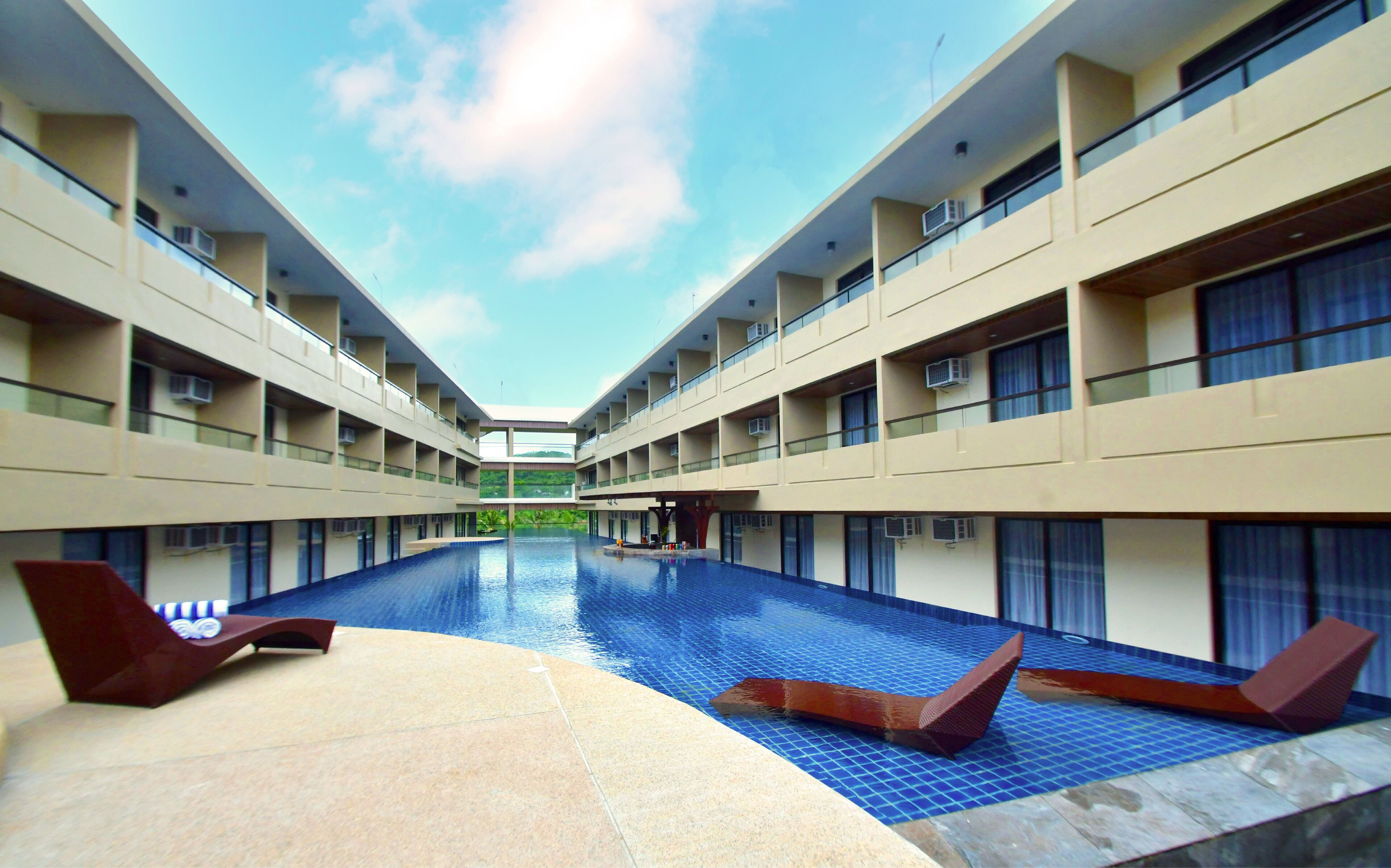 A modern hotel with a central swimming pool, multiple balconies, and a clear sky.