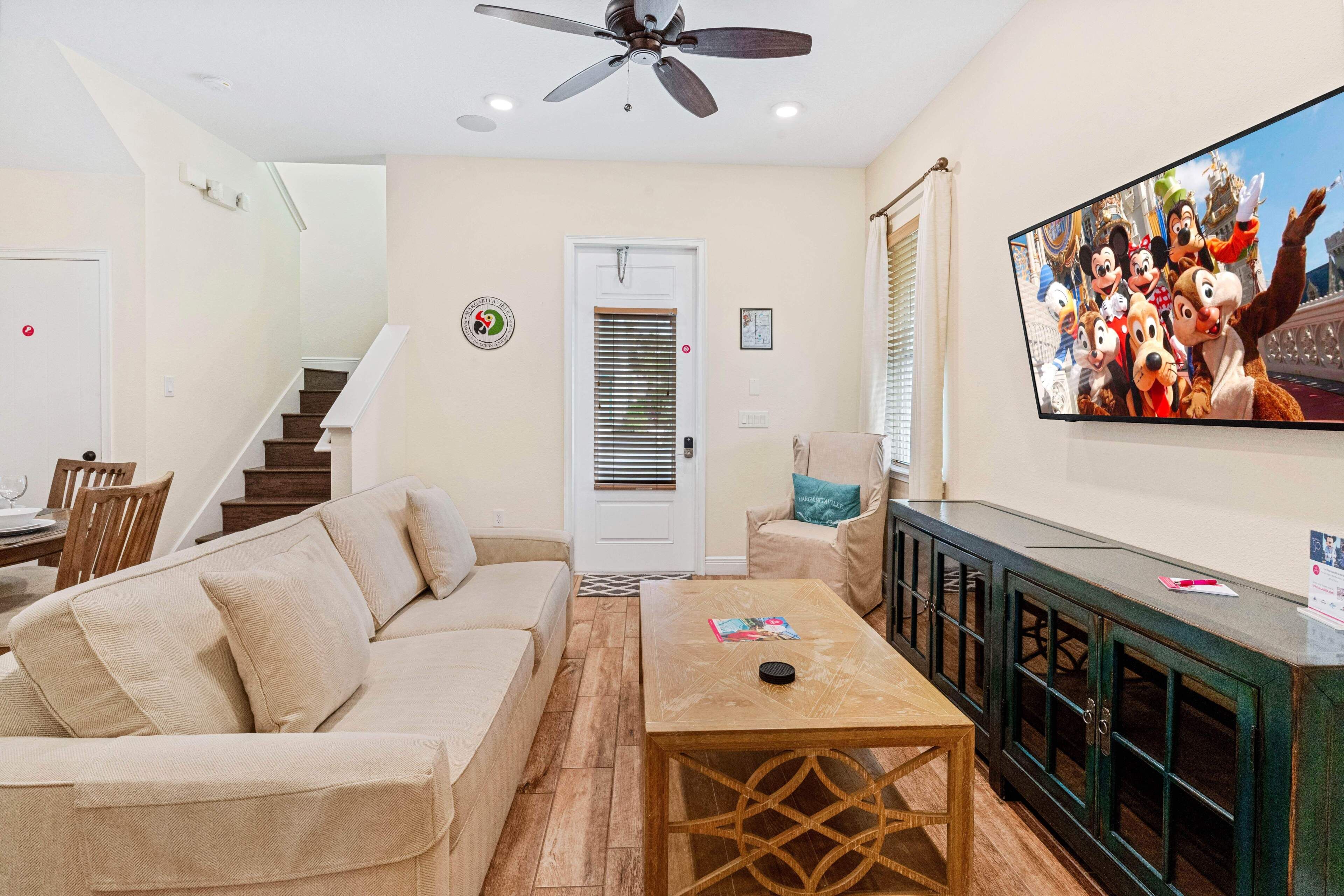 A living room with a beige sofa, a wooden coffee table, a television displaying animated characters, a staircase, and a door with a window.