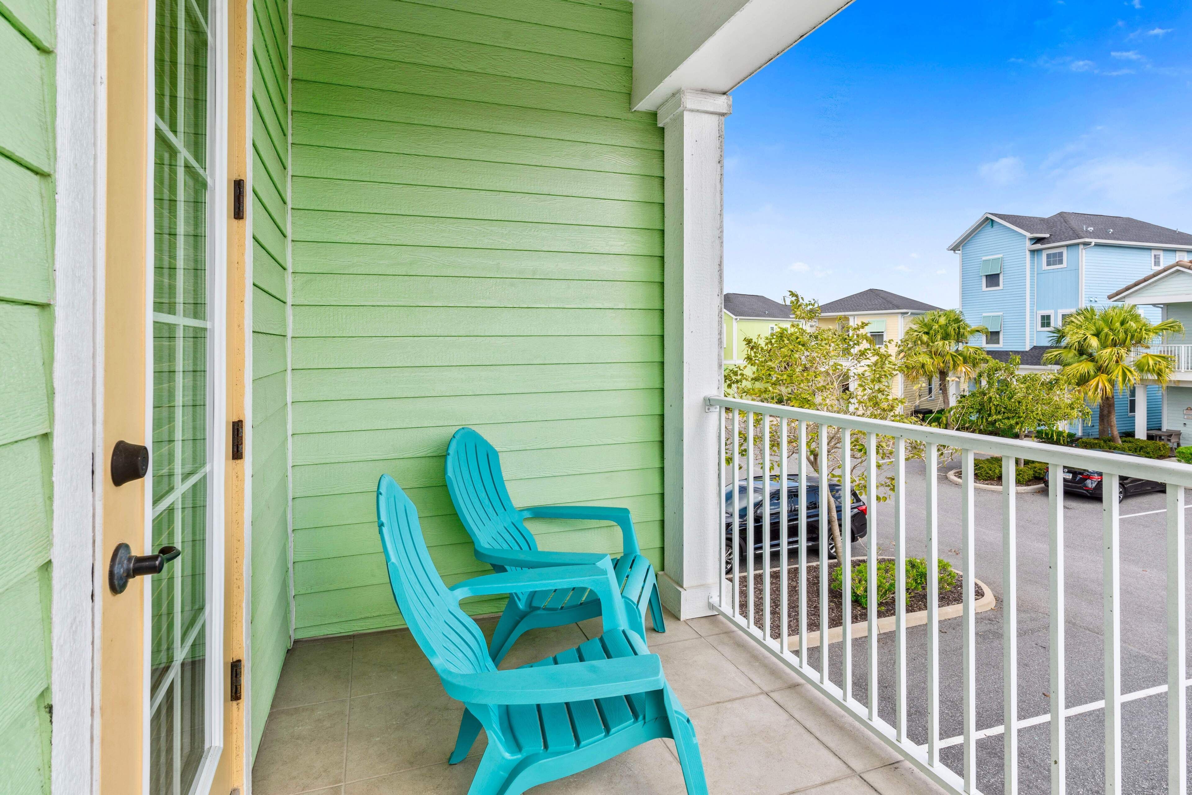A green building with a white balcony railing, two turquoise Adirondack chairs, and a view of neighboring houses.