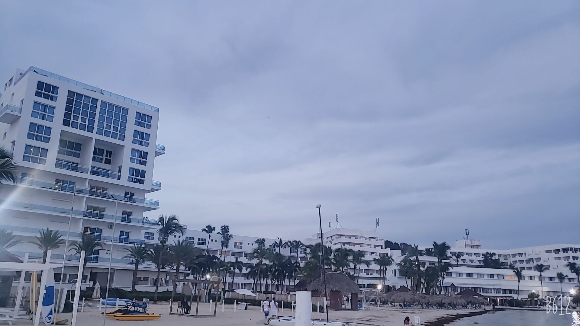 A beachfront hotel with multiple floors, palm trees, and a beach area with umbrellas and a lifeguard stand.