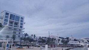 Un hotel frente a la playa con varios pisos, palmeras y una zona de playa con sombrillas y un puesto de socorristas.