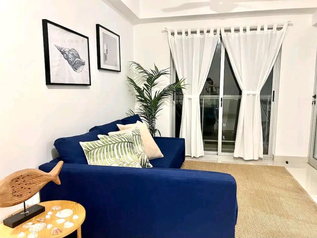 A living room with a blue sofa, a small round table with decorative items, a potted plant, and a large window with white curtains.