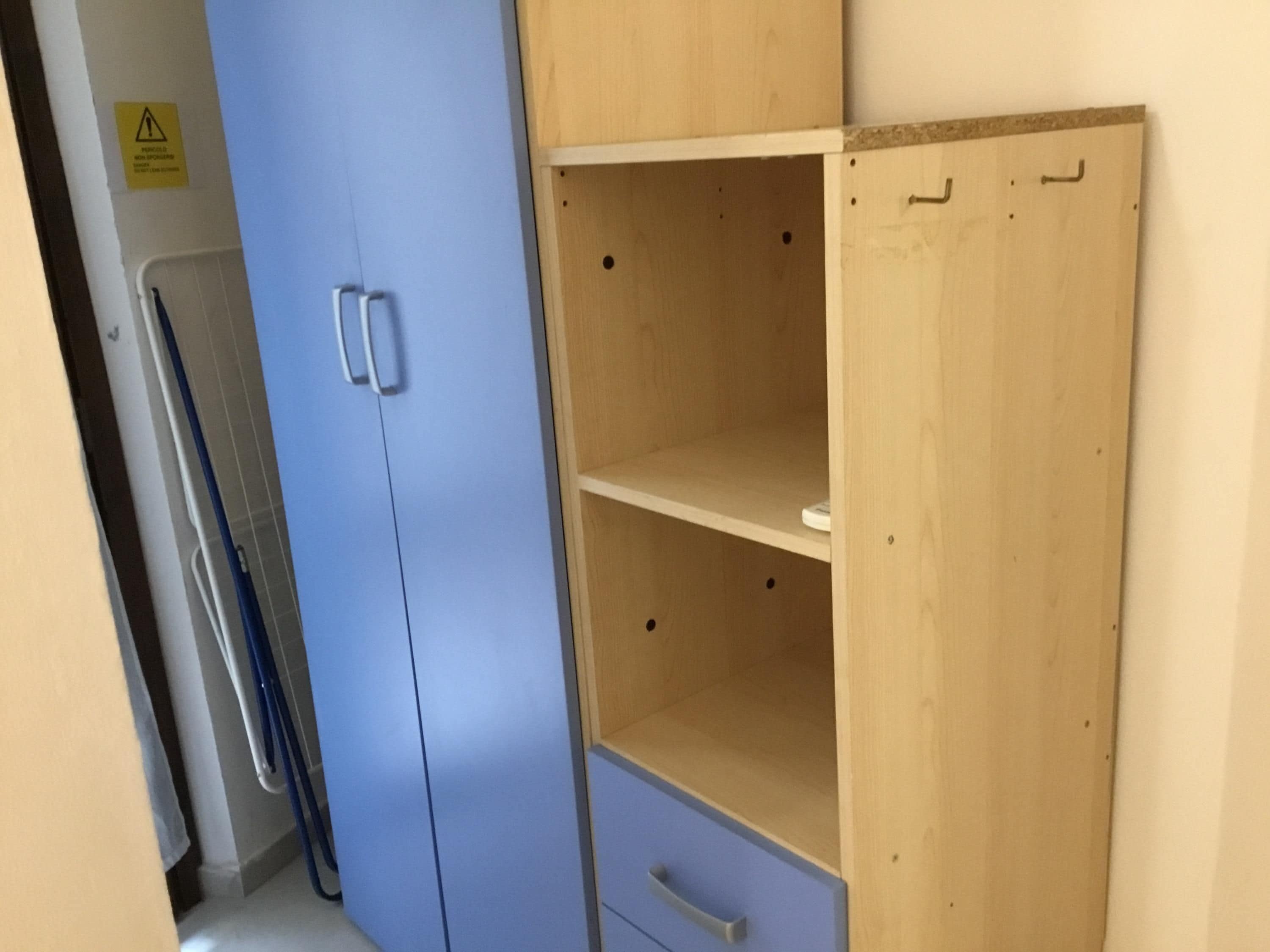 A room with a blue wardrobe, a wooden cabinet with shelves, and a white wall with a caution sign.