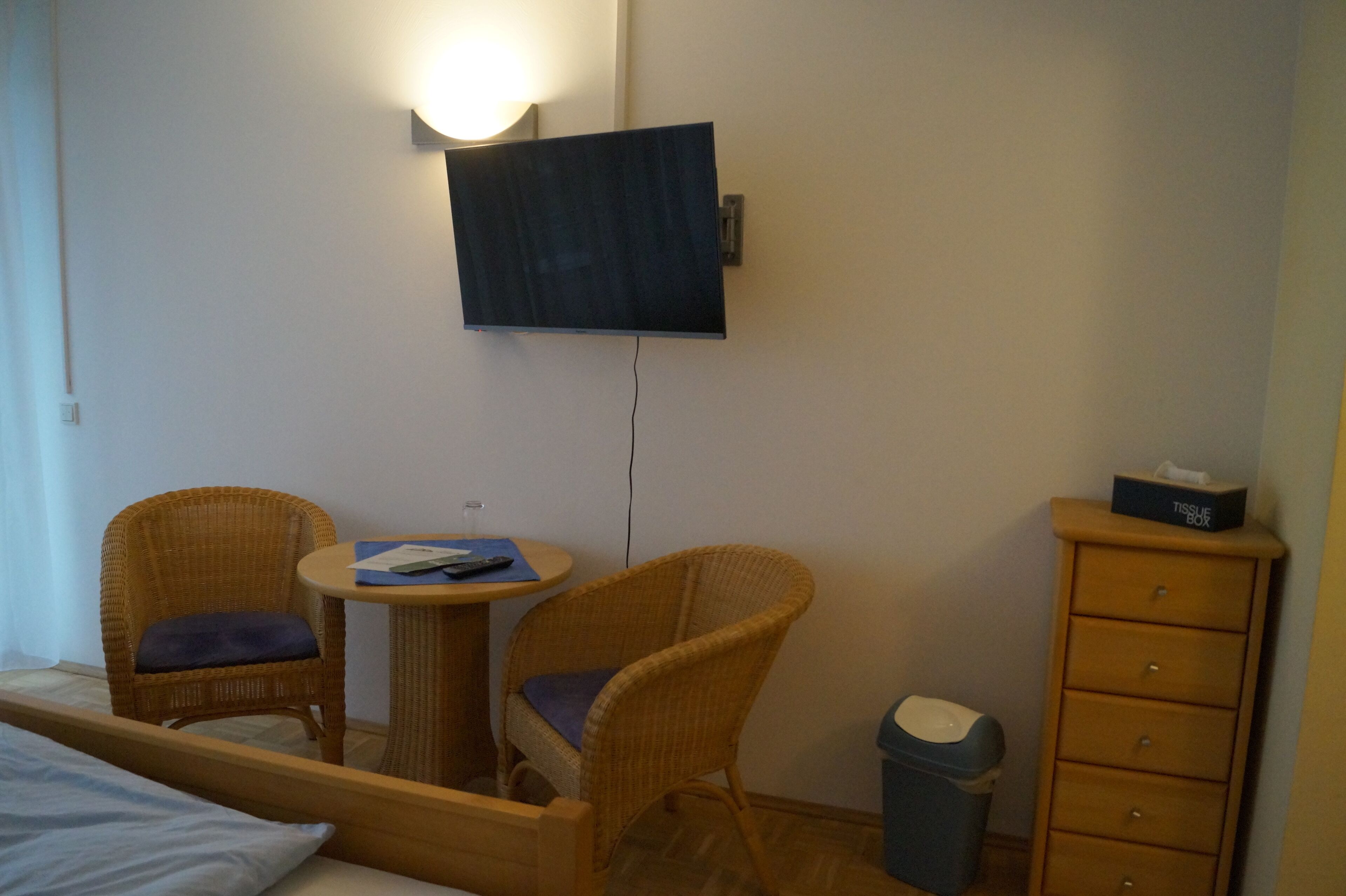Una habitación de hotel con una cama, dos sillones de mimbre, una mesita redonda, un televisor montado en la pared y un aparador de madera con un dispensador de pañuelos encima.