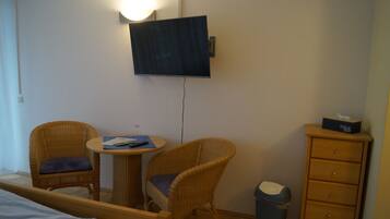 Una habitación de hotel con una cama, dos sillones de mimbre, una mesita redonda, un televisor montado en la pared y un aparador de madera con un dispensador de pañuelos encima.
