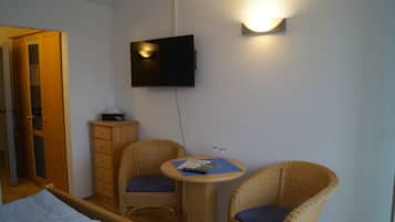 A room with a bed, two wicker chairs, a small table, a TV mounted on the wall, and a wooden cabinet.
